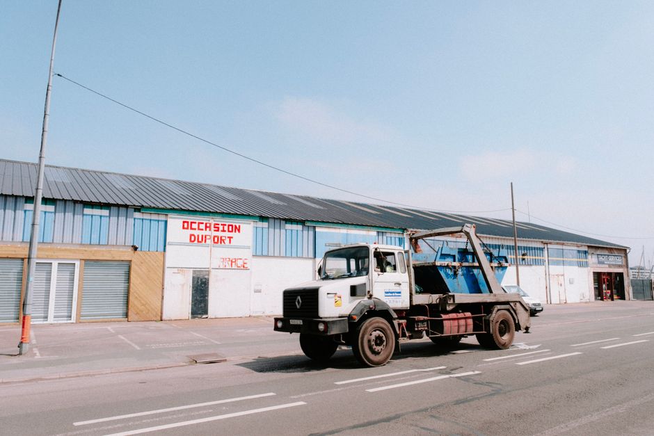 An industrial truck moves past a warehouse on a sunny day, highlighting urban transport.